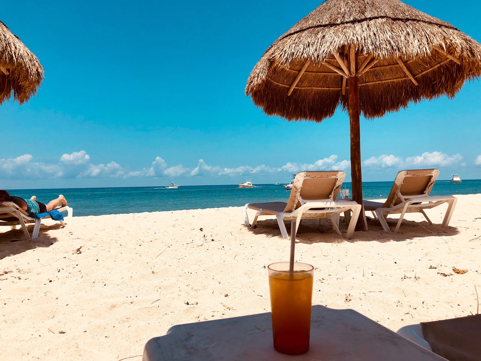 Palapa y camastros en una playa de arena blanca con una bebida en primer plano