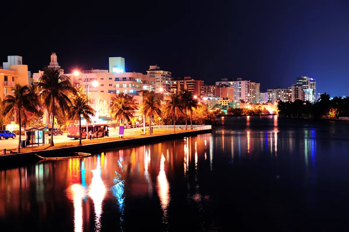 Malecón con edificios y palmeras iluminados reflejados en el agua de noche