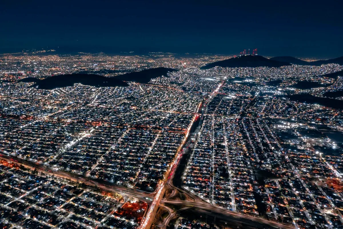 Vista aérea nocturna de una ciudad iluminada con avenidas y cerros al fondo