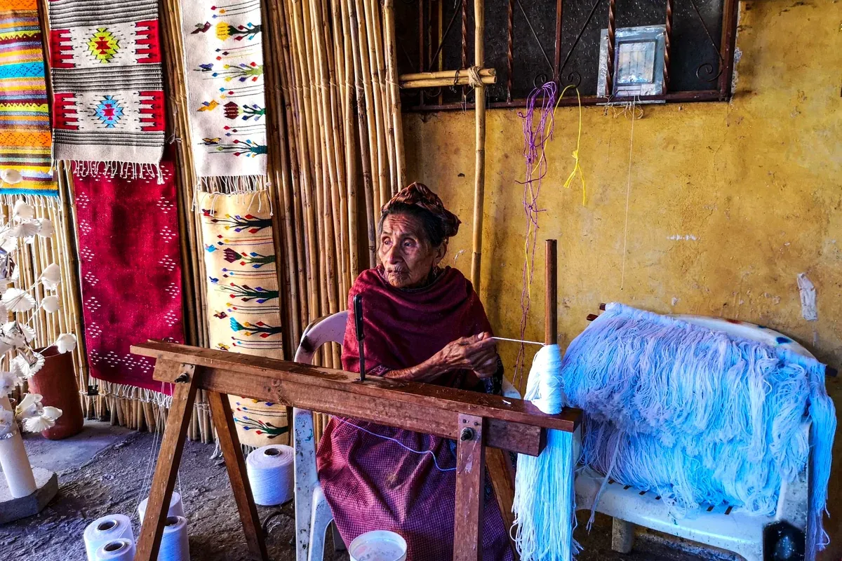 Tejedora en un telar de pedal rodeada de tapetes de colores