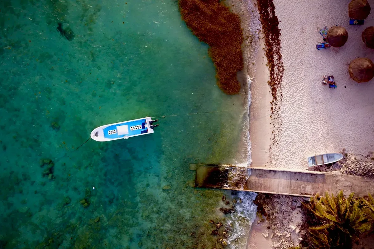 Lancha pequeña frente a una playa con palapas y un muelle de madera, desde el aire