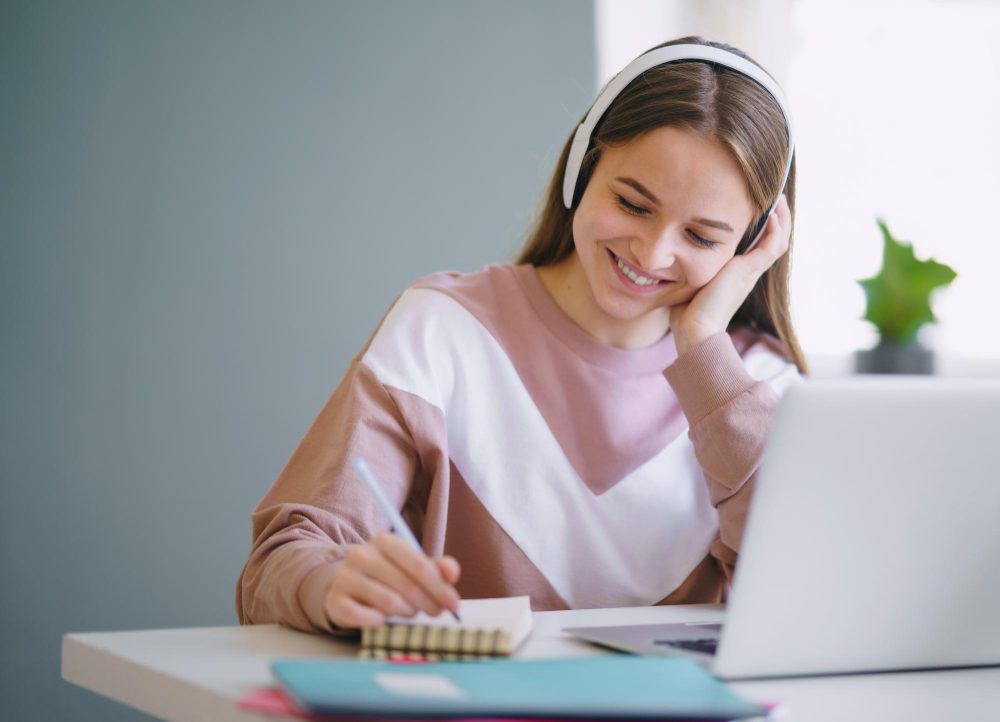 Joven sonriente con audífonos que escribe en un cuaderno frente a una laptop