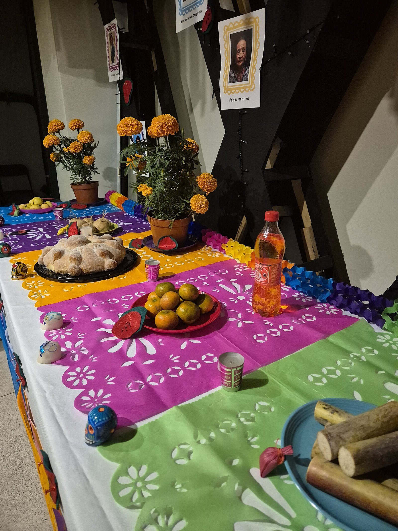 Mesa de ofrenda con pan de muerto, mandarinas, caña de azúcar, macetas de cempasúchil, calaveras de azúcar y una fotografía enmarcada