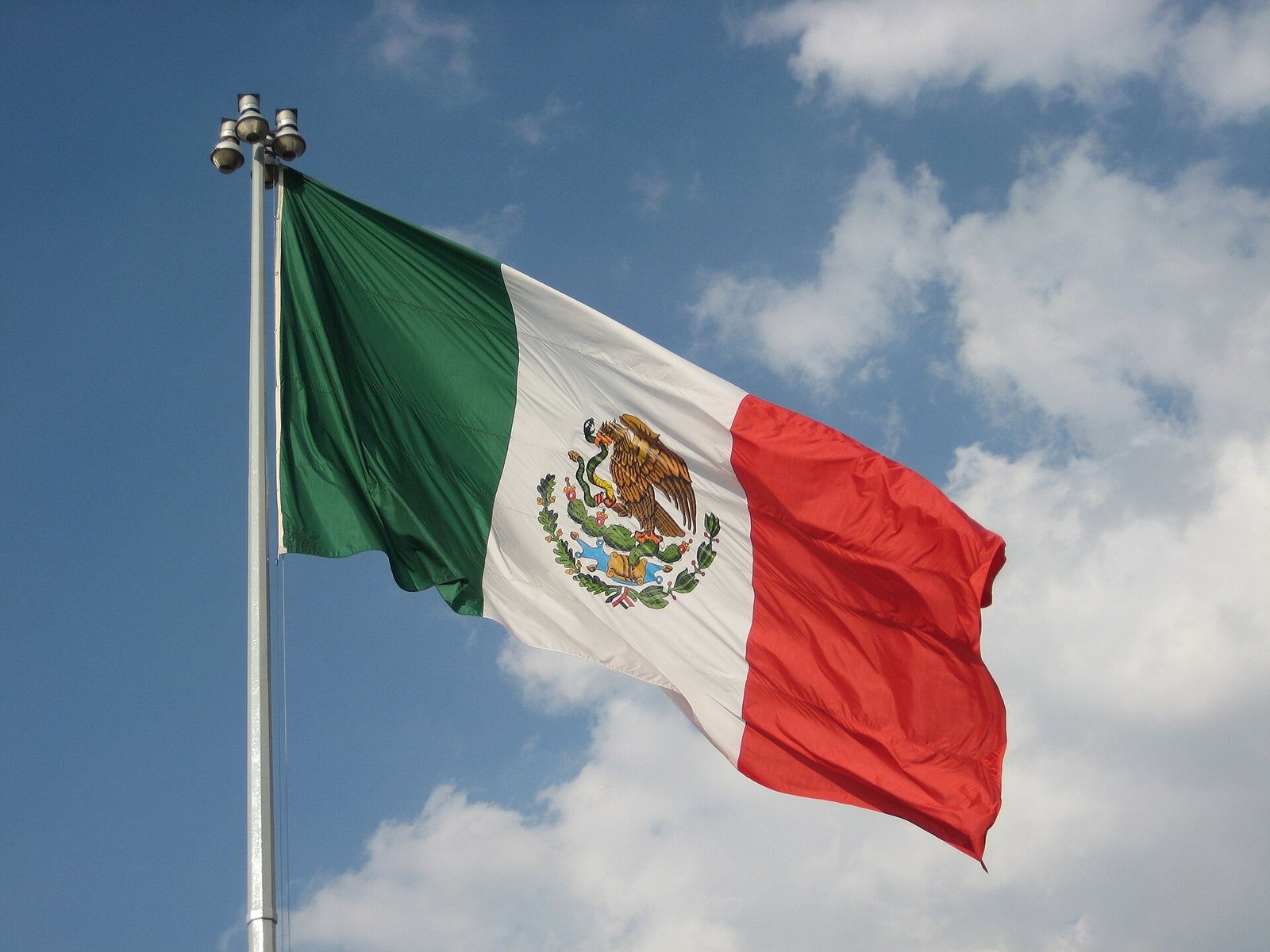 Bandera de México ondeando en lo alto de un asta con cielo azul y nubes