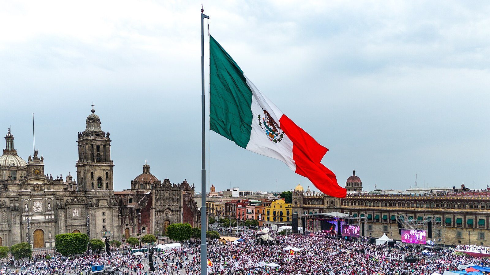 Bandera de México desplegada en el asta del Zócalo de la Ciudad de México, con la Catedral Metropolitana al fondo y la plaza llena de gente