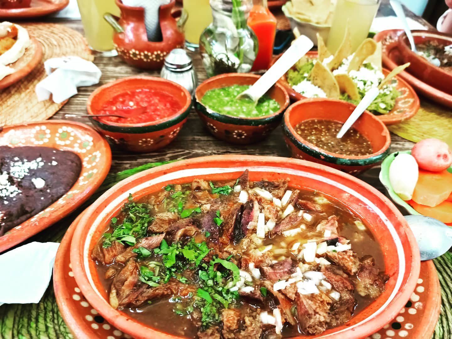 Cazuela de barro con birria de chivo con cilantro y cebolla, rodeada de salsas, frijoles y tacos