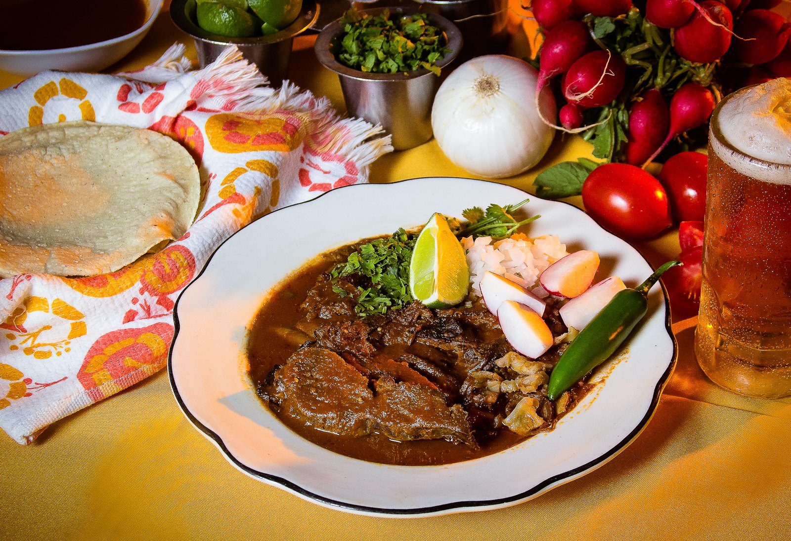 Plato de birria tatemada con cilantro, cebolla, limón y chile, junto a tortillas, rábanos y un vaso de bebida