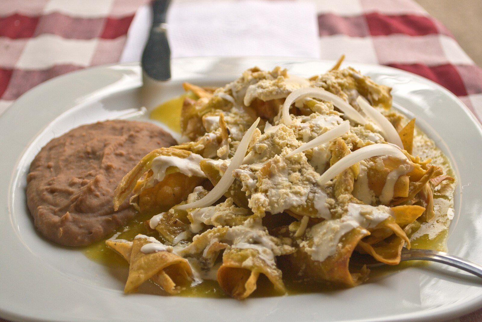 Chilaquiles verdes con crema y queso, acompañados de frijoles refritos, en plato blanco sobre mantel de cuadros