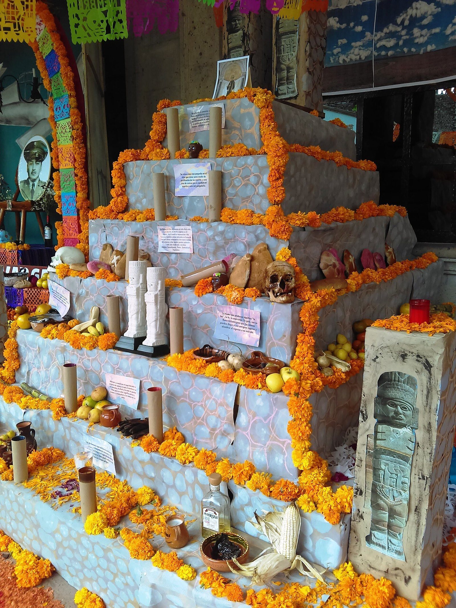 Altar escalonado cubierto de pétalos de cempasúchil, con velas, calaveras, frutas, papel picado y una figura de piedra