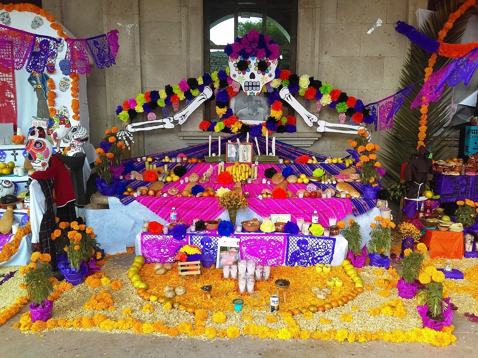 Altar de Día de Muertos de varios niveles con retrato de un hombre mayor, calaveras de colores, pan, frutas y arco de flores de cempasúchil