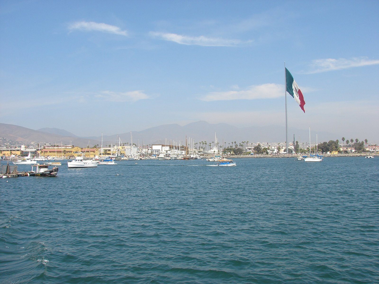 Enorme bandera de México ondeando en un asta sobre una bahía con veleros y lanchas, con cerros al fondo