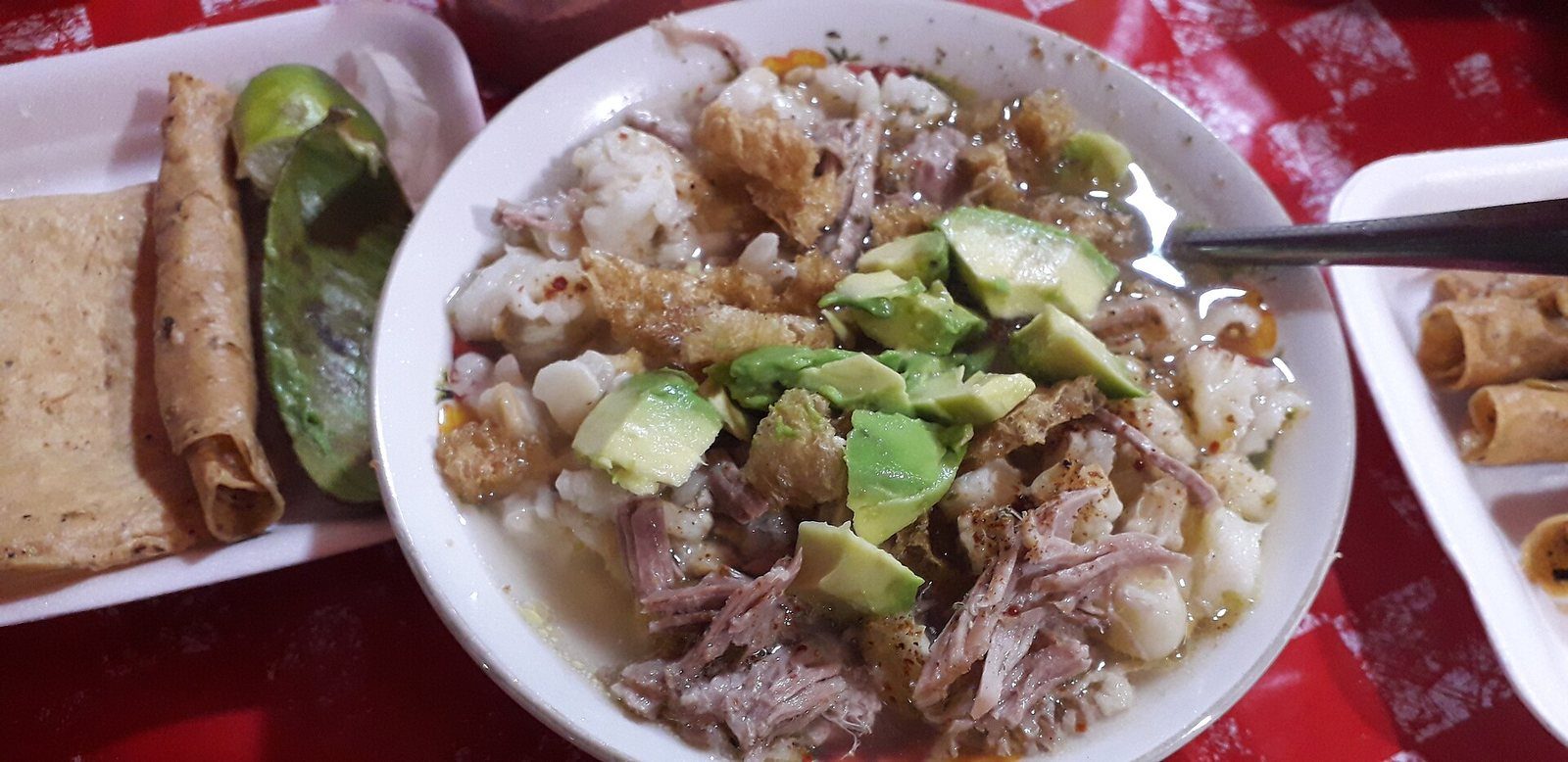 Plato de pozole blanco con carne deshebrada, chicharrón y rebanadas de aguacate, junto a tostadas y limones