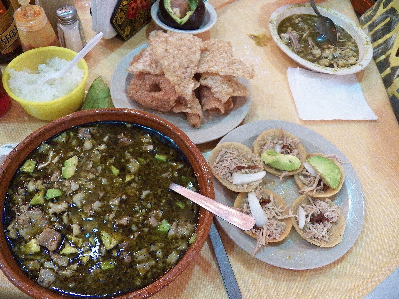 Cazuela de barro con pozole verde, junto a platos de chicharrón, tostadas con aguacate y cebolla picada
