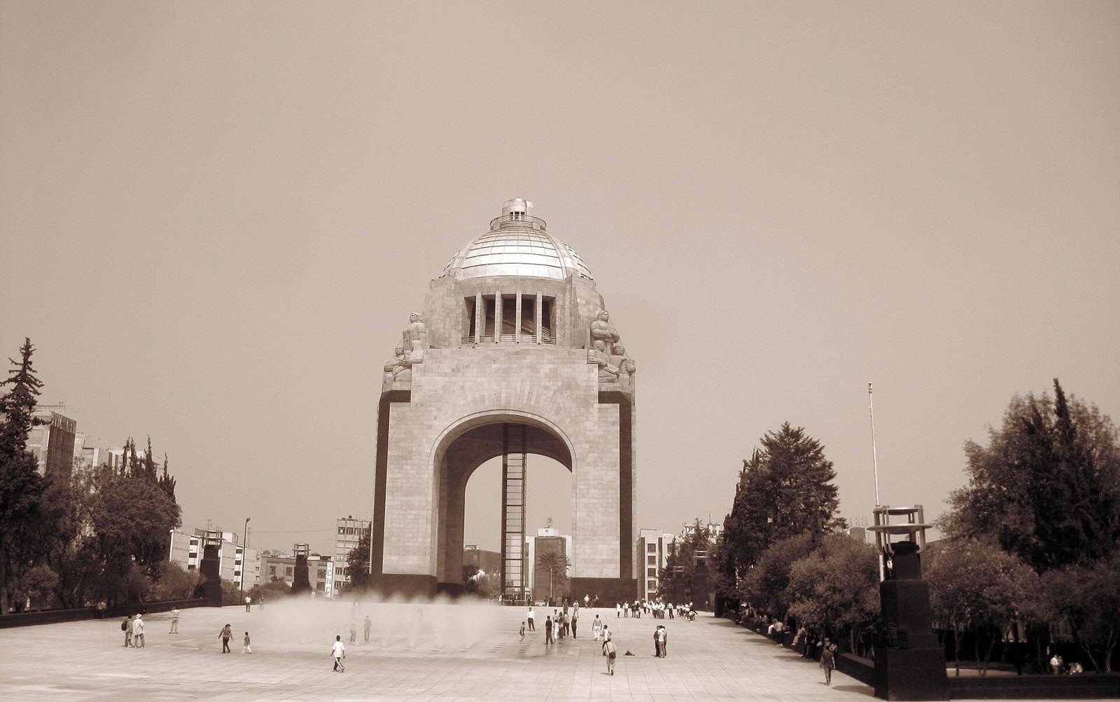 Vista amplia del Monumento a la Revolución en tono sepia, con su arco y cúpula al fondo de una explanada con visitantes y árboles